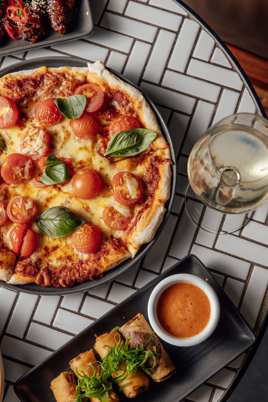 Pizza with cherry tomatoes and basil, spring rolls and a glass of white wine on a tiled table